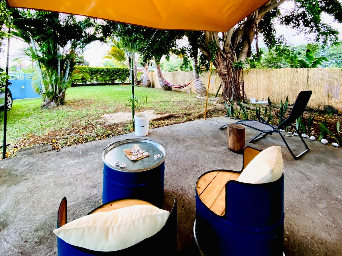 A shaded outdoor area is presented, featuring two blue barrel chairs with cushion seating. A small round table sits between them, and a hammock is visible in the background. Lush greenery surrounds the space, providing a serene environment.
