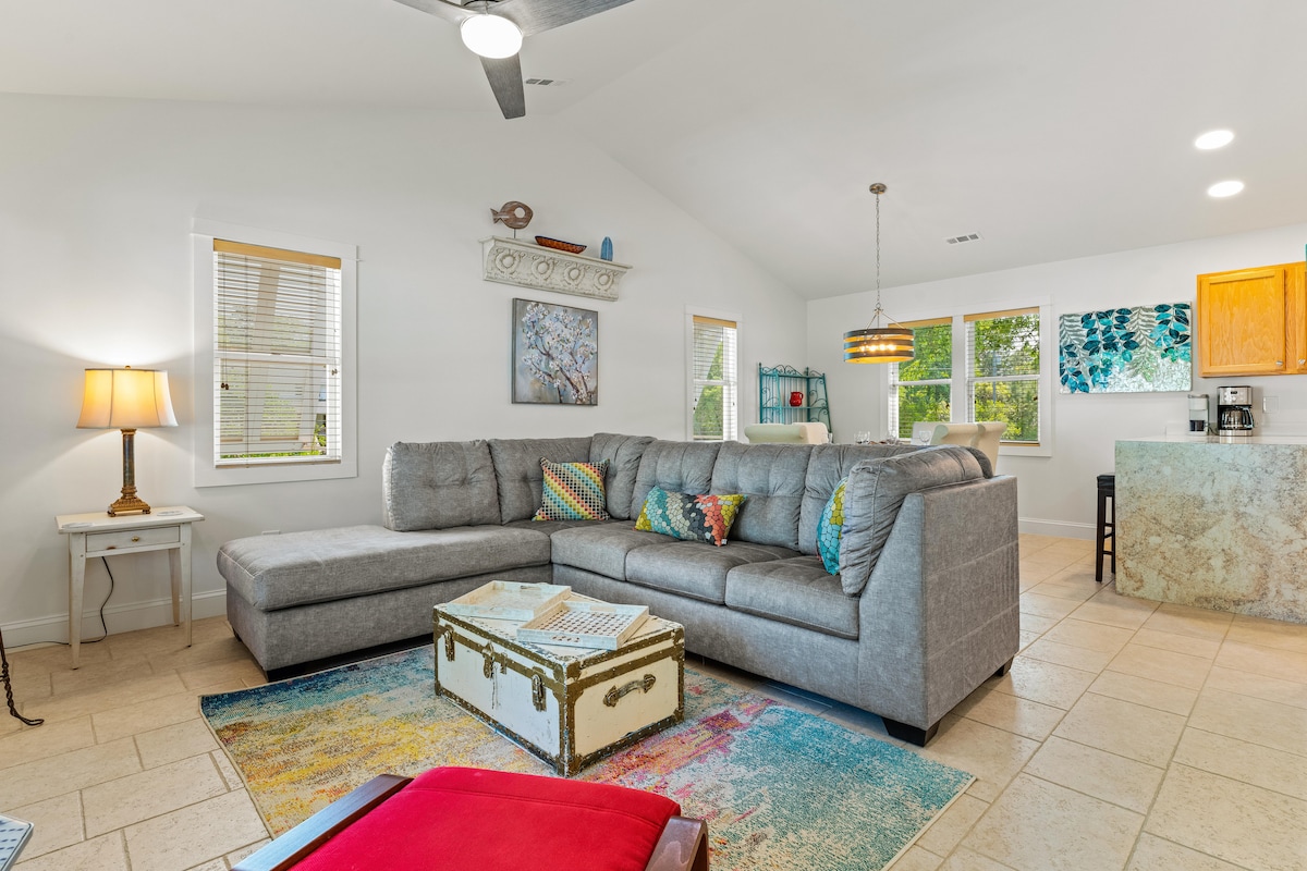 A spacious living area features a large sectional sofa adorned with colorful cushions. A glass coffee table sits atop a vibrant area rug. Natural light fills the room through multiple windows, highlighting the open layout that connects to the adjacent kitchen.