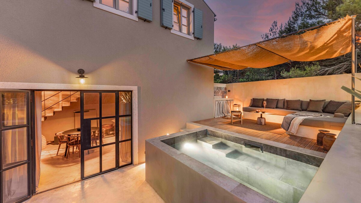 An outdoor living area features comfortable seating under a large canopy, alongside a small heated pool. Glass doors lead into the home, where a dining space and an inviting interior can be seen. Soft lighting creates a calming atmosphere during twilight.