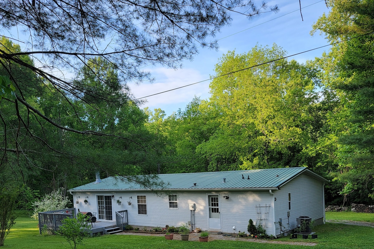 The exterior of the recently updated home is shown, with a green metal roof and surrounded by lush trees and greenery. A spacious deck is visible at the front, along with several flower planters lining the walkway.