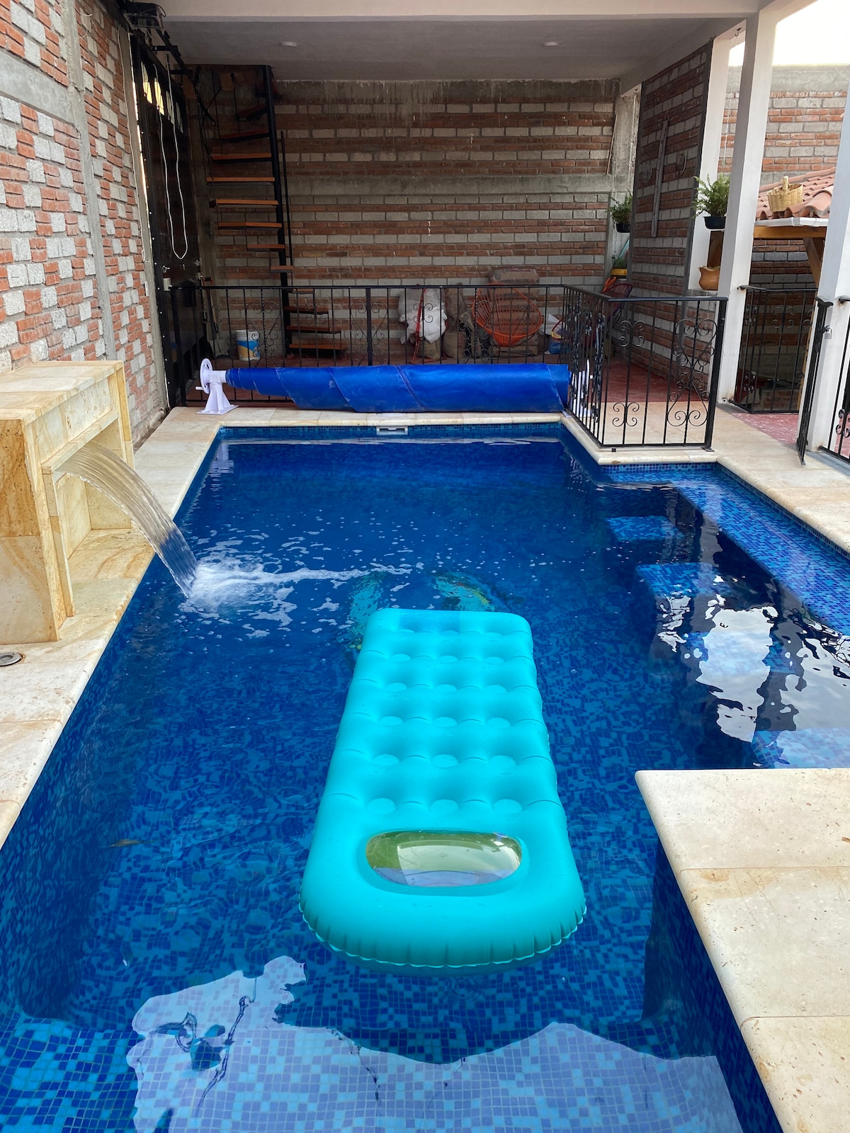 A swimming pool is featured, with a bright blue inflatable float resting on the water's surface. The pool area is surrounded by stone and tile, and includes a built-in fountain adding a gentle flow of water. Staircase and various loungers are visible in the background.