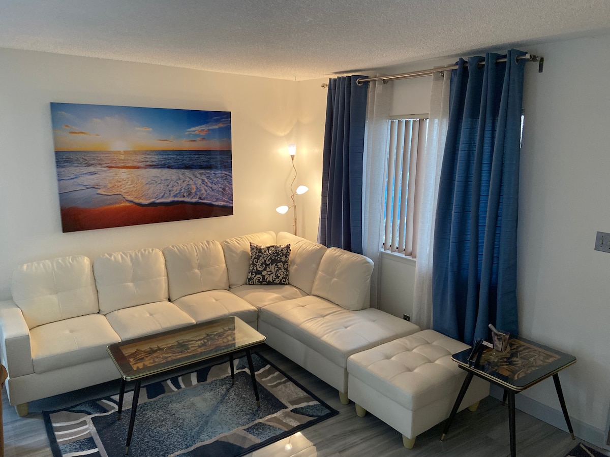 A bright living room features a large white sectional sofa with decorative pillows. Two elegant coffee tables are positioned on a patterned area rug. Natural light enters through vertical blinds, while blue curtains add a soft touch to the space. A landscape painting hangs on the wall.
