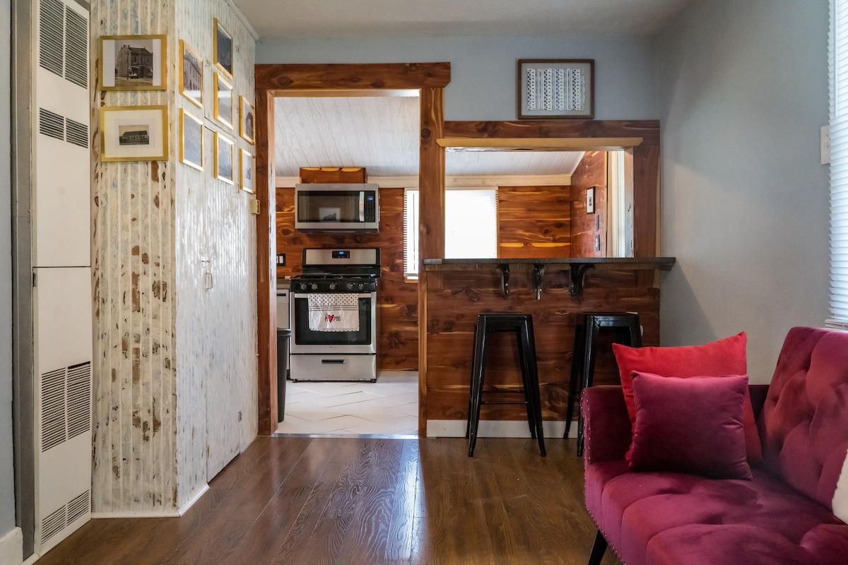 A cozy interior showcases a plush seating area with a bold red couch, leading to a kitchen area with wooden paneling. Stainless steel appliances and granite countertops are visible. Three sleek black bar stools are situated at the counter, contributing to a welcoming atmosphere.