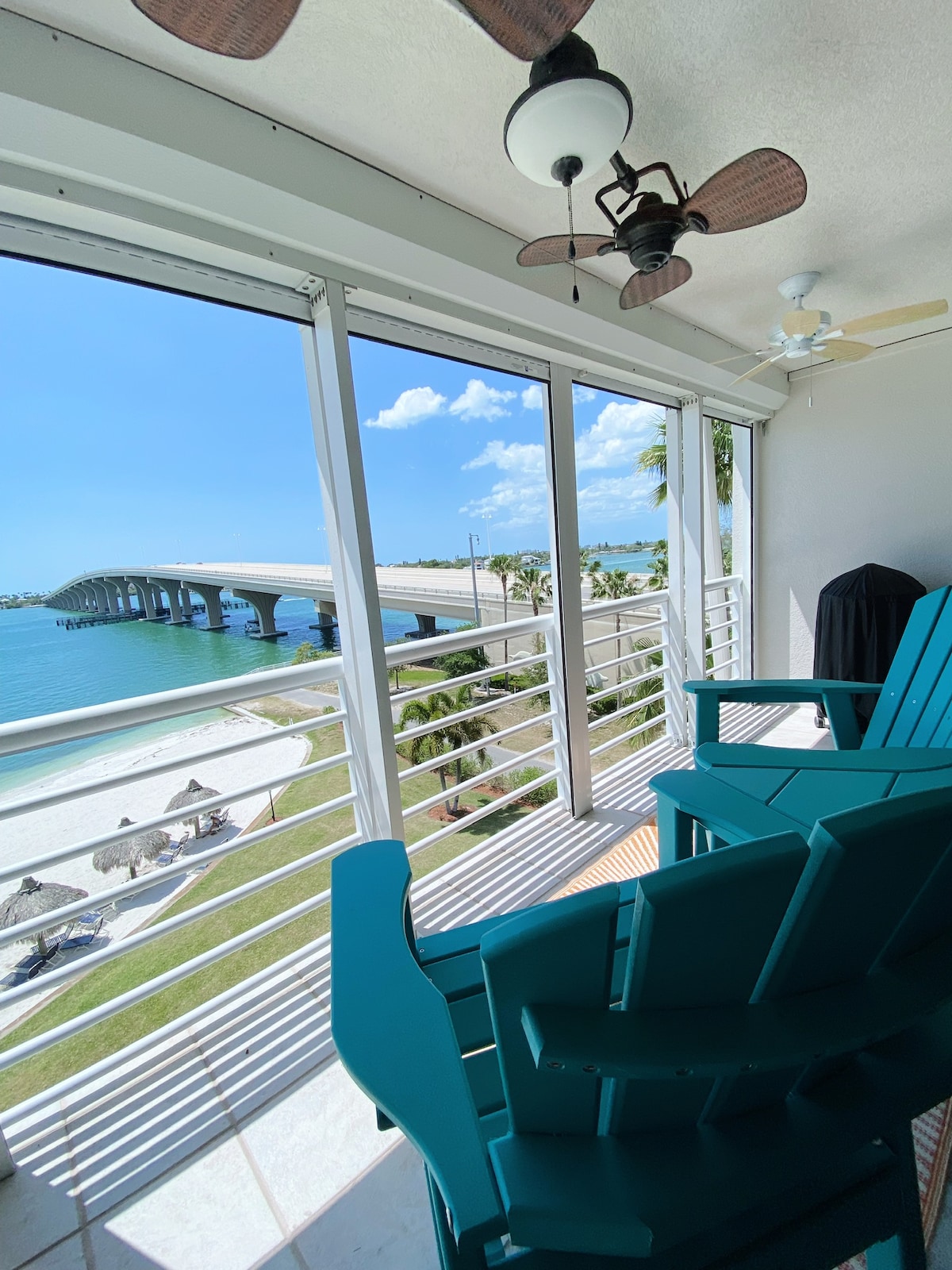 A spacious balcony features comfortable turquoise chairs overlooking a tranquil beach and water. In the distance, a bridge is visible against a bright sky, enhancing the scenic view. The open design allows for ample natural light and fresh air.