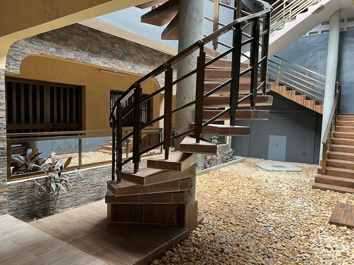 A winding staircase with wooden steps and a black railing draws the eye upward, surrounded by a serene courtyard featuring smooth stones and minimal plant life. The architectural design incorporates natural light, enhancing the open space's tranquility.