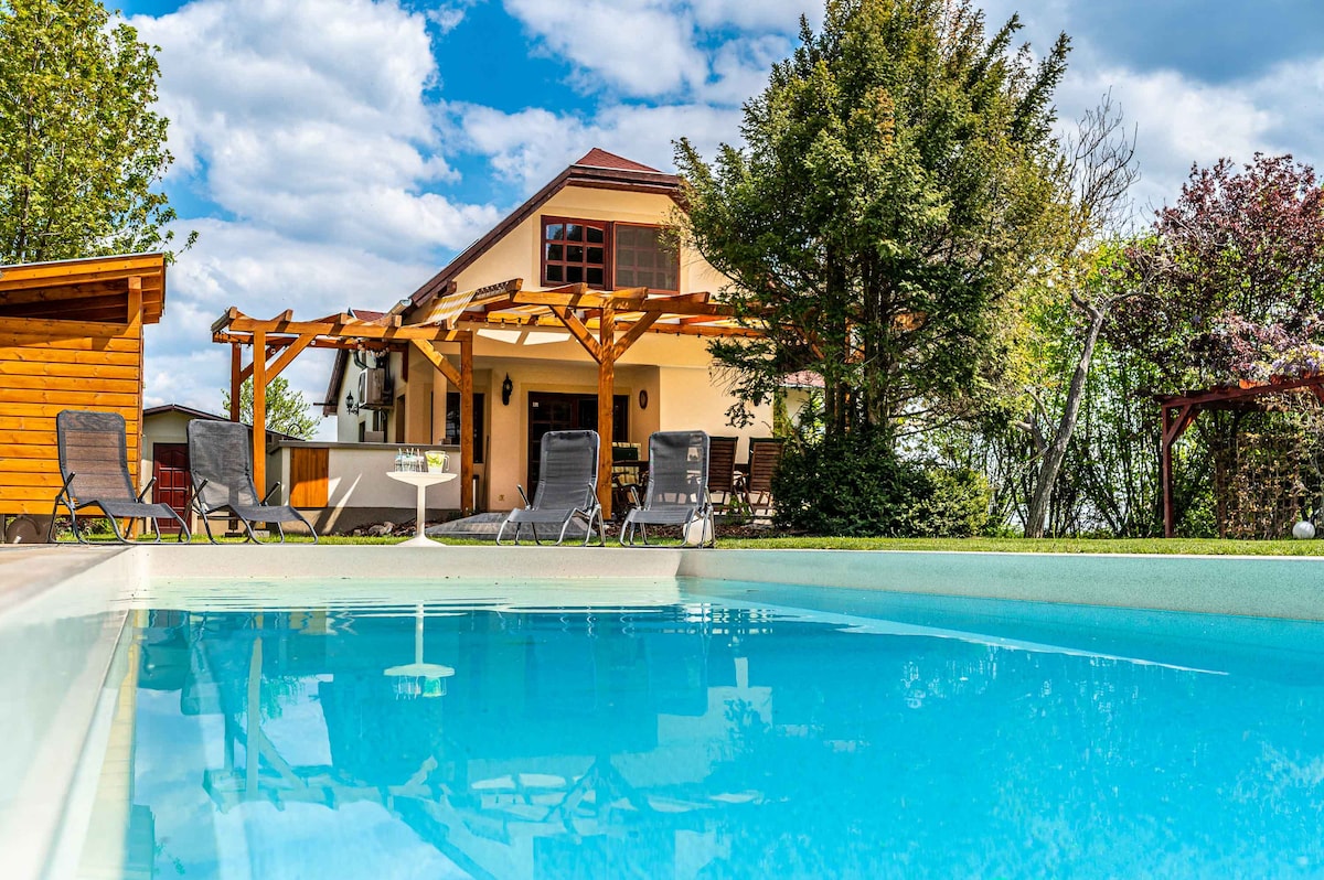 A clear view of a swimming pool reflects the sky and surroundings, with lounge chairs positioned around it. The house features a classic design with red accents, surrounded by lush greenery and wooden structures, including a pergola for outdoor seating.