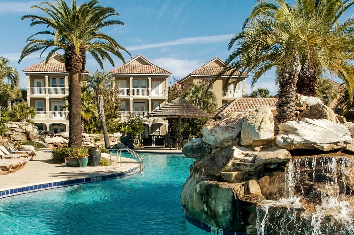 A lagoon-style pool with a rock waterfall is surrounded by palm trees and tropical landscaping. In the background, a two-story building is visible, featuring balconies and a tiled roof, creating a serene resort environment.