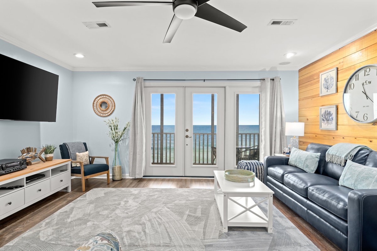 A bright living area showcases a navy blue sofa adorned with decorative pillows. A large flat-screen TV is mounted on the wall opposite. Floor-to-ceiling glass doors provide an unobstructed view of the ocean, while a plush area rug enhances comfort underfoot.