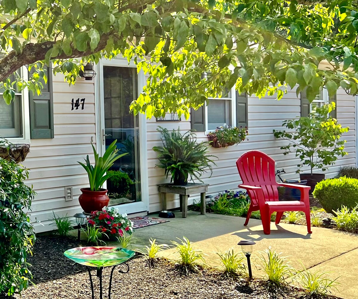 A welcoming entrance is framed by lush greenery and flowering plants. A bright red chair on the patio invites relaxation, while decorative garden elements are visible around the entryway. Sunlight filters through the leaves, creating a warm and inviting atmosphere.