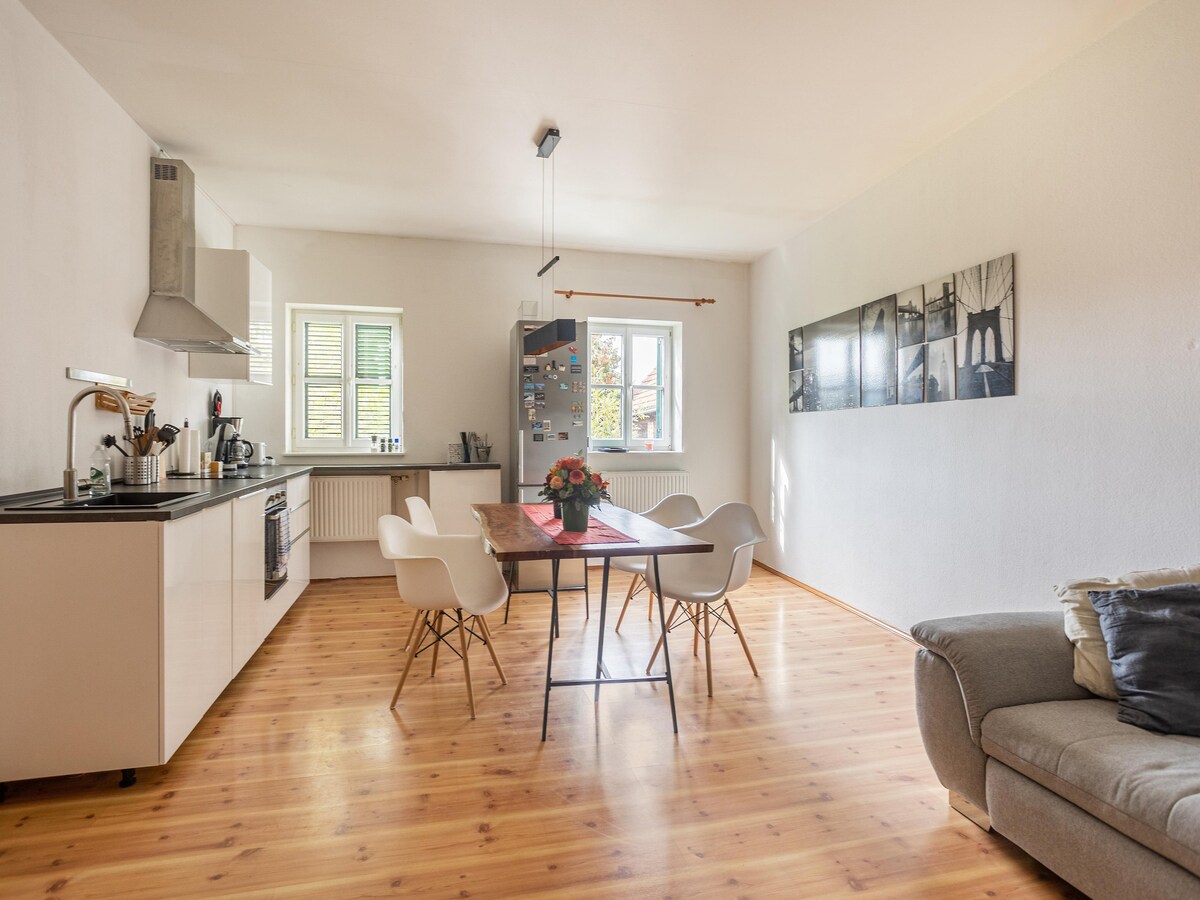 A modern kitchen area is highlighted by stylish furnishings, including a wooden dining table surrounded by white chairs. A comfortable sofa is positioned nearby. Natural light spills through multiple windows, enhancing the bright and airy ambiance.