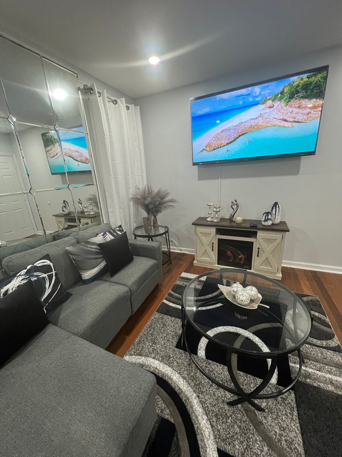 A modern living area features a comfortable gray sectional sofa, accented with decorative pillows. A sleek glass coffee table sits in front, displaying ornamental decor. The room is illuminated by soft overhead lighting and a large TV mounted on the wall showcases a serene beach scene.