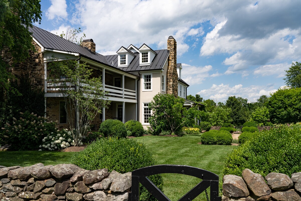 A charming countryside estate is framed by lush greenery and manicured gardens. The two-story home features multiple gables and a covered porch. A stone wall outlines the property, adding rustic character to the scenic setting.