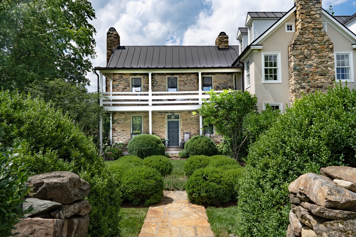 The rustic stone facade of the home is framed by lush greenery and well-maintained gardens. A stone pathway leads to the inviting entrance, with a covered balcony on the second level and large windows allowing natural light to fill the space.