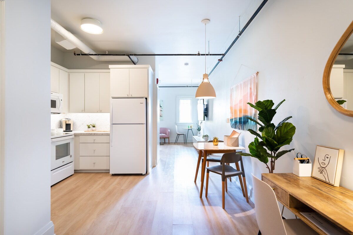 A spacious kitchen space features bright white cabinetry and appliances, with a dining table for two situated nearby. A dedicated workspace is positioned against the wall, complemented by a potted plant. Warm, natural light filters through a nearby window.