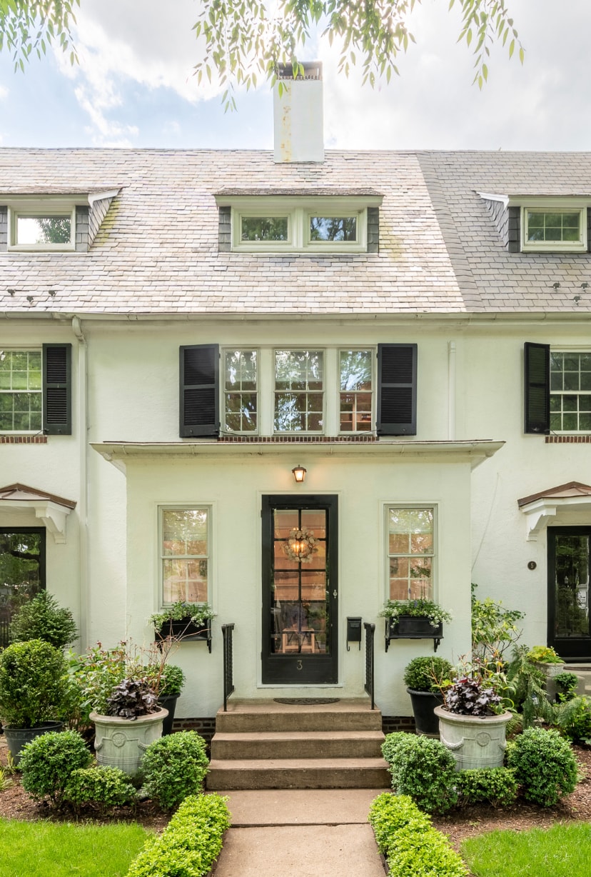 The exterior of a charming townhouse presents a well-maintained façade with large windows framed by black shutters. A welcoming front door is flanked by decorative potted plants. Lush greenery lines the walkway leading up to the entrance, enhancing the inviting atmosphere of the home.