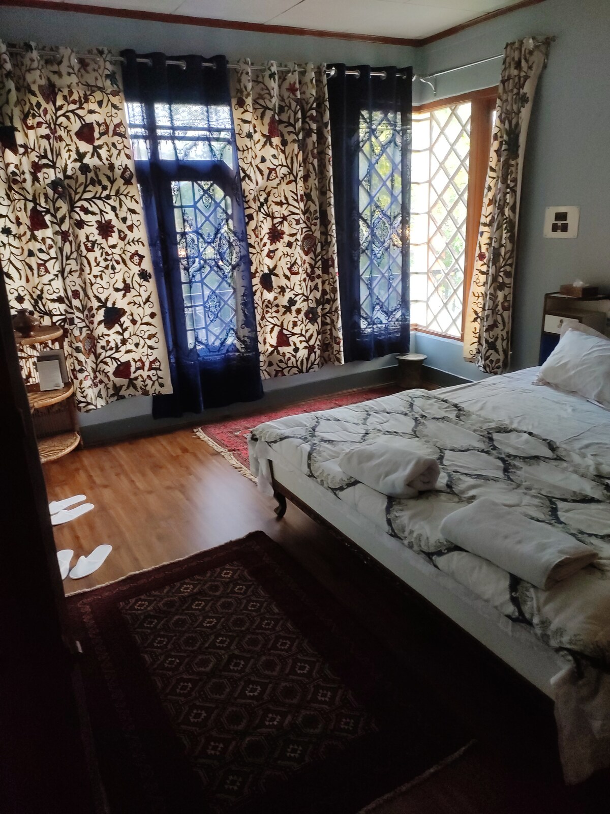 A comfortable bedroom is furnished with a spacious bed adorned with white bedding. Elegant curtains with floral patterns frame the windows, allowing soft light to filter in. A patterned rug lies on the floor, complemented by a small bedside table and decorative items.