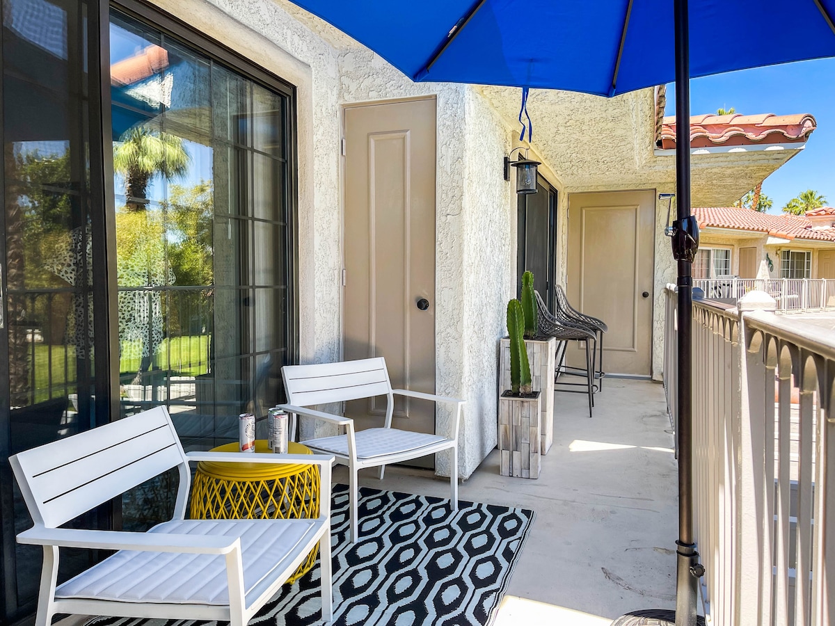 A patio area is furnished with two white chairs and a yellow accent table, situated on a patterned rug. A blue umbrella provides shade, while a potted cactus adds a natural touch. The setting offers views of the surrounding greenery and nearby buildings.
