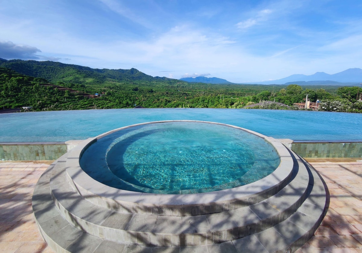 An inviting infinity pool with a circular jacuzzi is visible at the forefront, surrounded by stone steps. Lush green hills and distant mountains create a serene backdrop, under a clear blue sky, reflecting a tranquil atmosphere.