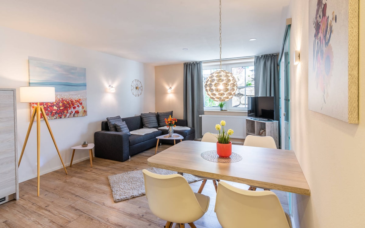 A modern living space is presented with a comfortable gray sofa and a decorative coffee table. A dining table with white chairs is set beneath a stylish light fixture. Bright artwork adorns the walls, and natural light flows through a window, enhancing the inviting atmosphere.