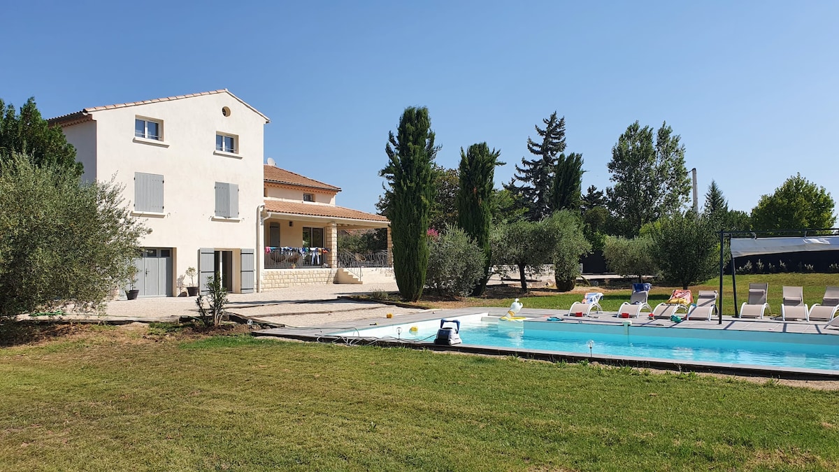 A spacious renovated house is set on a large lawn, surrounded by trees. A private swimming pool is visible, bordered by several sun loungers. The exterior features a covered terrace that extends from the house, providing shaded outdoor space.