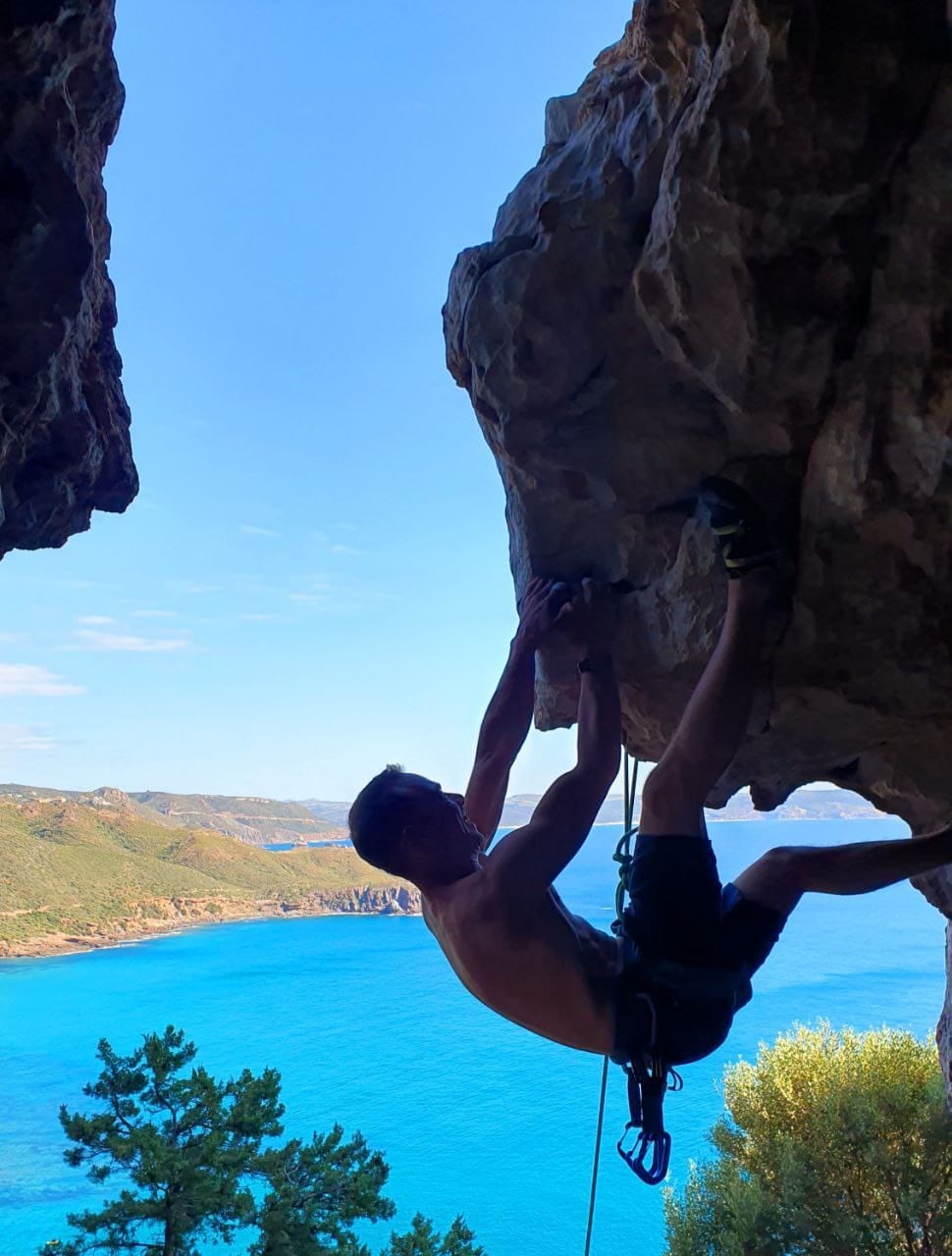 A climber is seen scaling a rocky cliff with the ocean view in the background. The blue water contrasts with the greenery of the surrounding hills, creating a vibrant landscape. The clear sky enhances the scenery, showcasing a serene outdoor environment ideal for climbing enthusiasts.