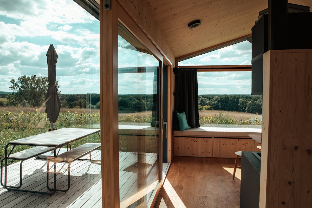 A cabin interior with large glass doors provides views of the surrounding greenery. A spacious wooden table and chairs are positioned on a deck, while a cozy seating area is placed by a window with natural light filtering in.