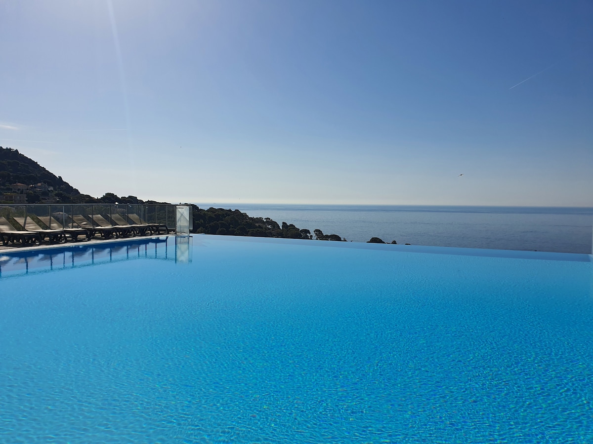 An infinity pool stretches toward the horizon, featuring bright blue water that meets the sky. Sun loungers line the edge, offering a perfect view of the sea, creating a serene environment for relaxation.