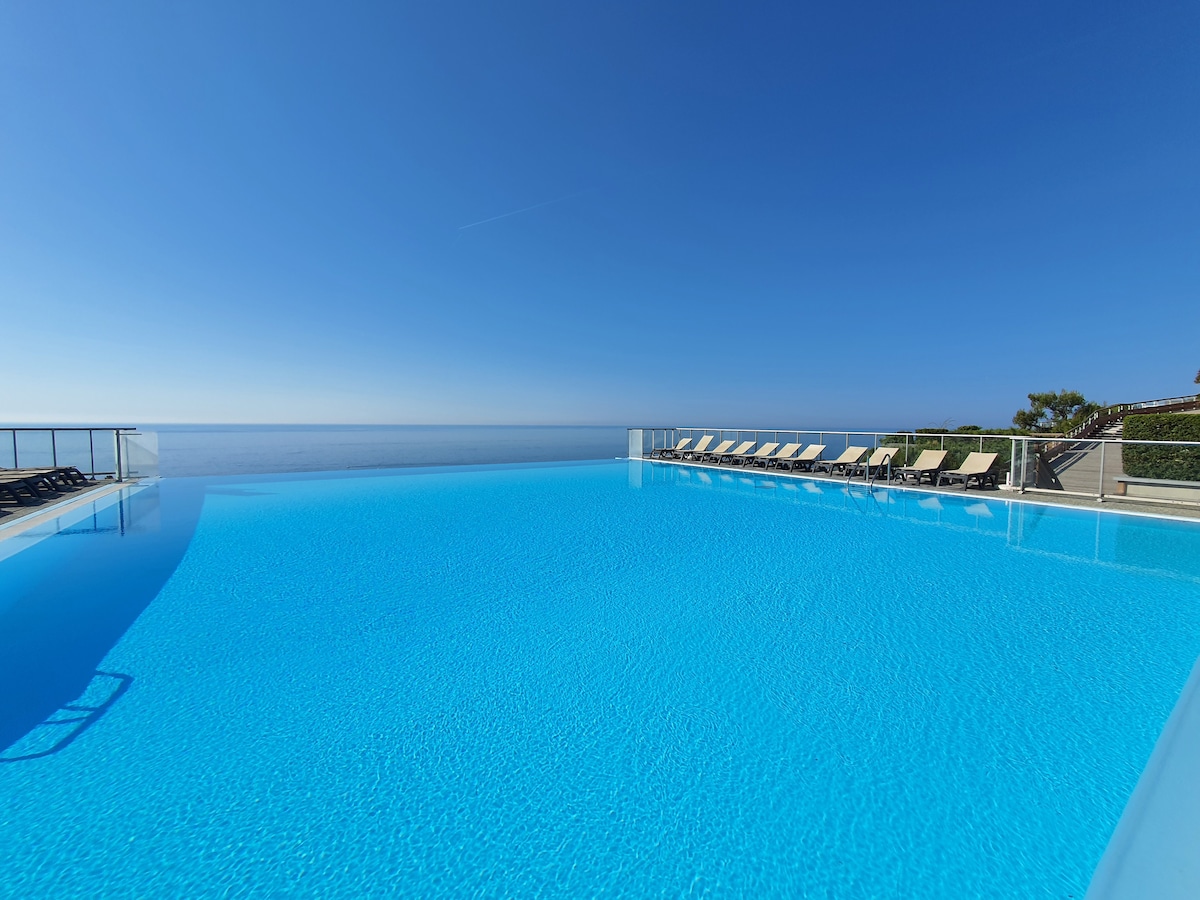 A modern infinity pool reflects the clear blue sky, offering a calming view over the landscape. Sun loungers are arranged neatly along the poolside, providing a relaxing space for guests to enjoy the serene environment.