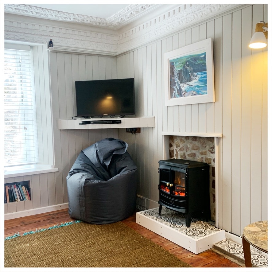 The lounge area is equipped with a flat-screen TV mounted on the wall, above a decorative stone fireplace. A cozy bean bag chair and a small side table are positioned nearby, while natural light filters in through the window, highlighting the textured wall paneling.