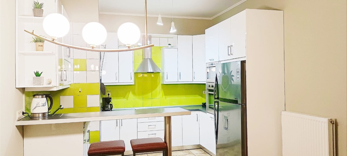 A modern kitchen features sleek white cabinetry and vibrant green backsplash. Stainless steel appliances, including a refrigerator and stove, are prominently displayed. A contemporary light fixture hangs above a small dining area with two brown stools, offering an inviting space for casual meals.