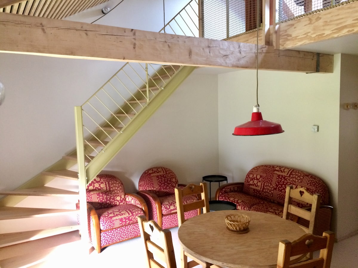 The interior features a warm seating area with two red-patterned sofas and a round wooden table. A wooden staircase leads to the mezzanine, with a pendant light hanging above. Natural light fills the space, emphasizing the inviting atmosphere.