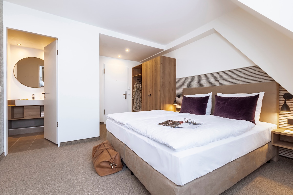 A spacious bedroom features a large bed with plush pillows and a neutral-colored comforter. Wooden cabinets provide storage space. The room is brightened by natural light coming through a nearby window, while a modern bathroom area is visible through an open door.