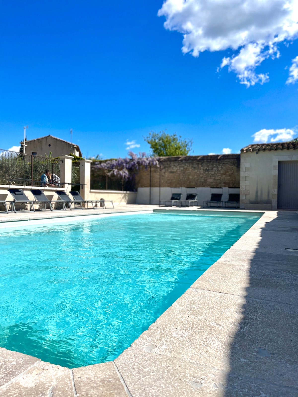 A shared pool area features a clear blue water pool surrounded by a spacious stone deck. Several lounge chairs are positioned along the edge, with a backdrop of greenery and low stone walls. A bright blue sky with scattered clouds enhances the inviting ambiance.