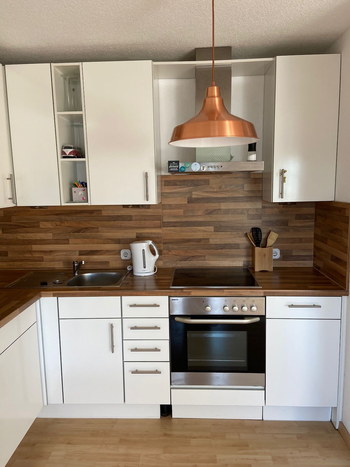 The kitchen features modern cabinetry in white and wood tones, accented by a copper pendant light. A compact layout includes a sink, oven, and stove with multiple drawers and shelves for storage. Essential appliances, such as a kettle, are visible.
