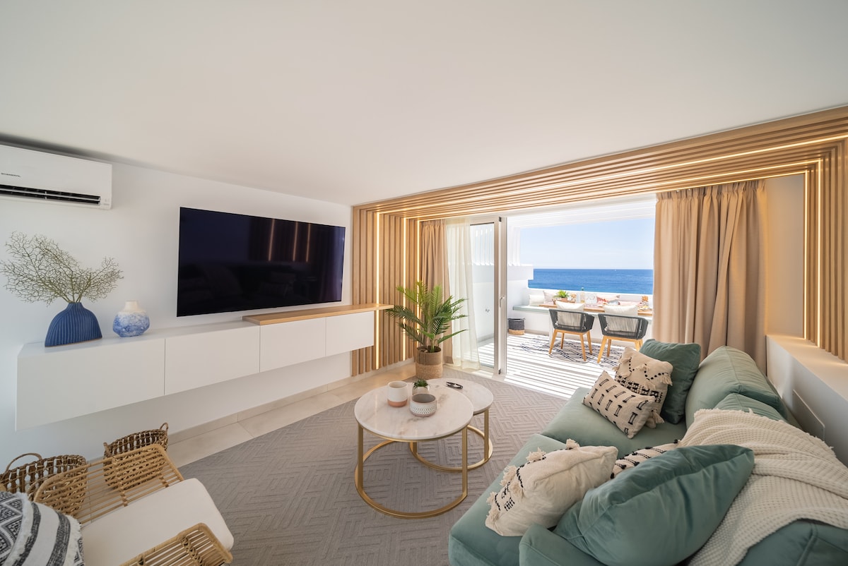 A light-filled living area features a comfortable sectional sofa, a round coffee table, and a large wall-mounted TV. Flowing natural light illuminates the space, while glass doors lead to a terrace showcasing ocean views. Tasteful decor, including a plant and decorative items, enhances the ambiance.