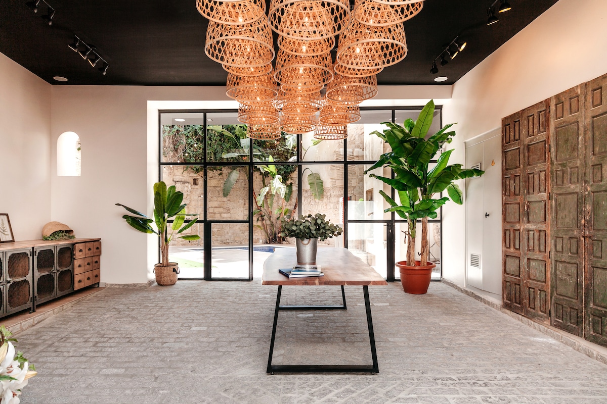 A spacious lobby area features large glass doors that provide views of the garden and pool area. A wooden table is centered beneath artistic woven light fixtures, while tall plants add a touch of greenery to the setting. Decorative wooden panels enhance the contemporary decor.