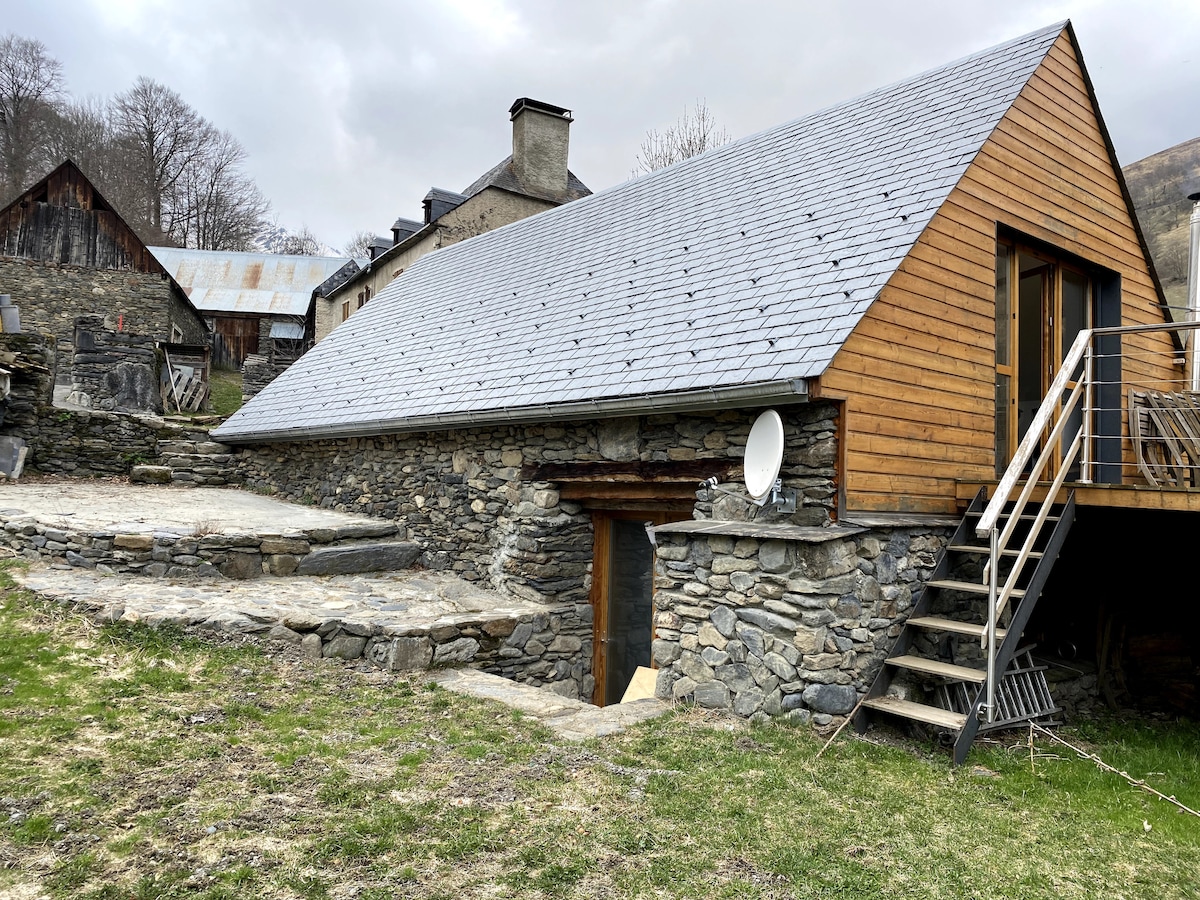 A rustic exterior features a mix of stone and wooden materials, highlighting the house's unique architecture. Steps lead to a raised entrance, while a stone terrace surrounds the lower level, creating a blend of natural textures. The surrounding landscape shows signs of early spring.