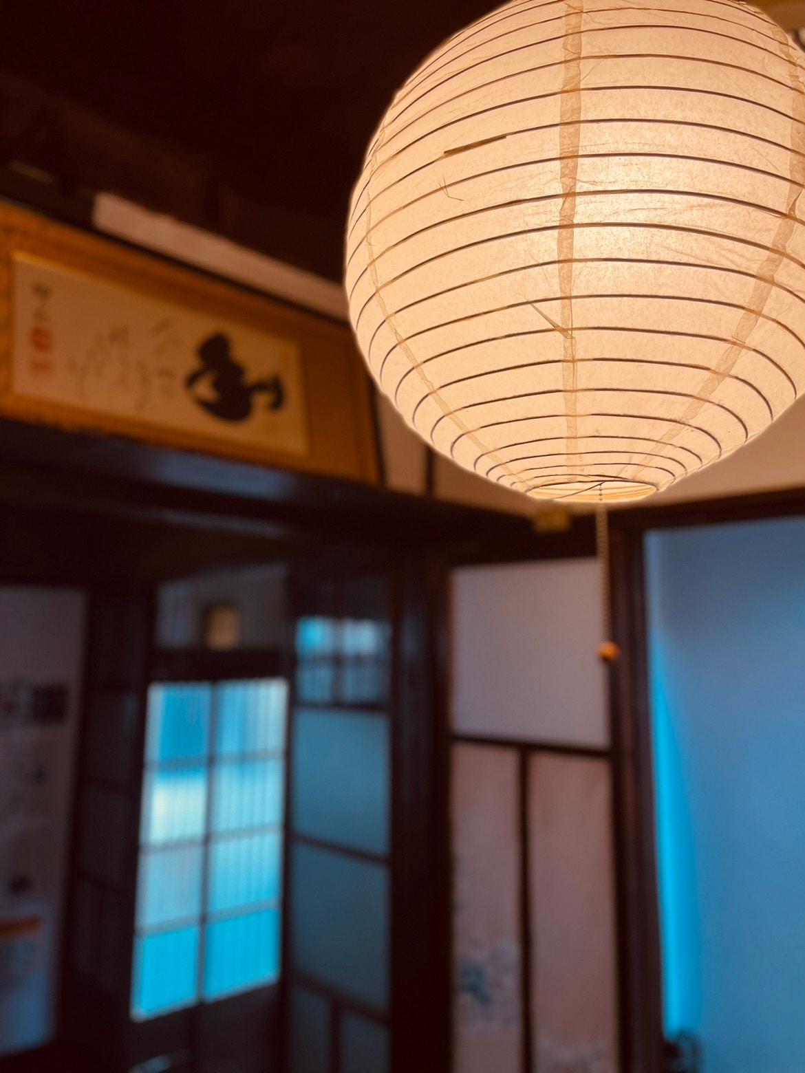 A traditional paper lantern hangs from the ceiling, softly illuminating the entryway. Sliding shoji screens are visible in the background, separated by wooden frames, contributing to the classic design of the space, with light seeping through to create a serene ambiance.