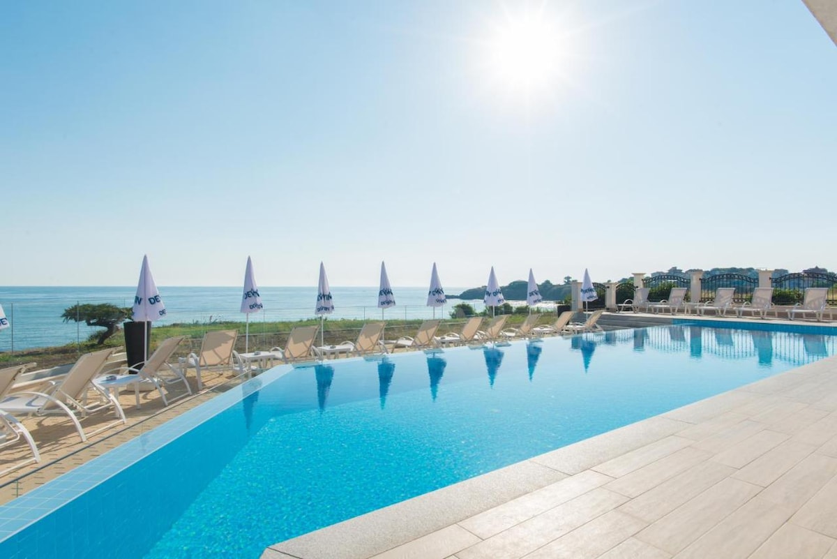 An infinity pool reflects sunlight, with lounge chairs arranged along its edge. White umbrellas provide shade while a clear view of the sea and beachfront is visible in the background, promoting relaxation in a serene environment.