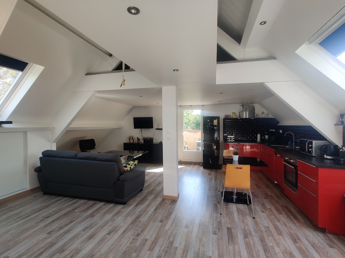A spacious open-concept living area features a well-equipped kitchen with red cabinetry. Natural light enters through skylights, illuminating the modern furnishings, including a comfortable gray sofa and a dining space with a wooden chair. A doorway leads to an outdoor terrace.