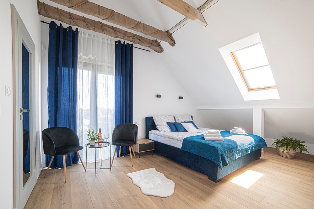 A bright and airy bedroom features a spacious bed adorned with blue and white linens. Two black chairs surround a small table, positioned near a window that allows natural light to fill the room. Wooden beams along the ceiling add a touch of warmth.
