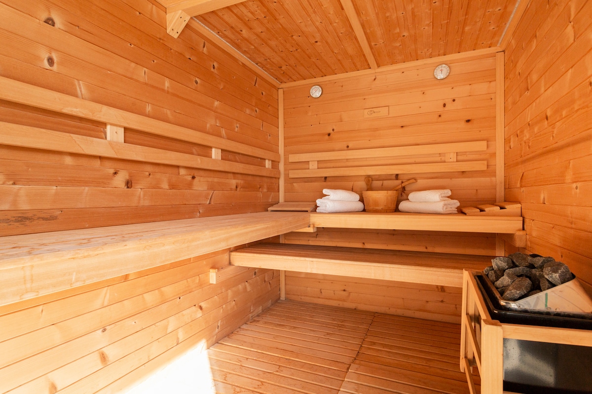 A wooden sauna features smooth wooden walls and a slatted bench, providing a warm and relaxing environment. Fresh towels are neatly arranged on the bench, and a decorative bowl is visible at the center. The sauna is well-lit with soft illumination, enhancing the natural wood aesthetic.