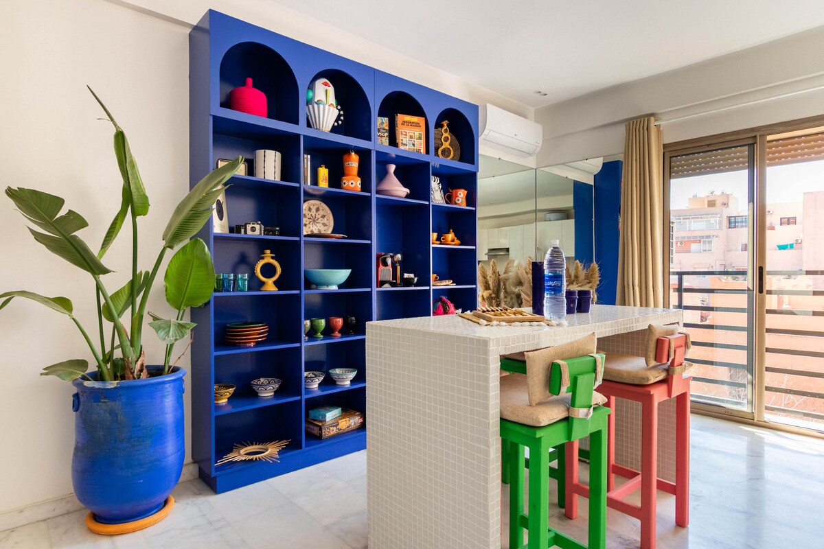 A vibrant dining area features a bright blue library showcasing an assortment of colorful decorative items and tableware. A modern table with bar stools sits at the forefront, while large windows allow natural light to fill the space, offering a view of the outside.