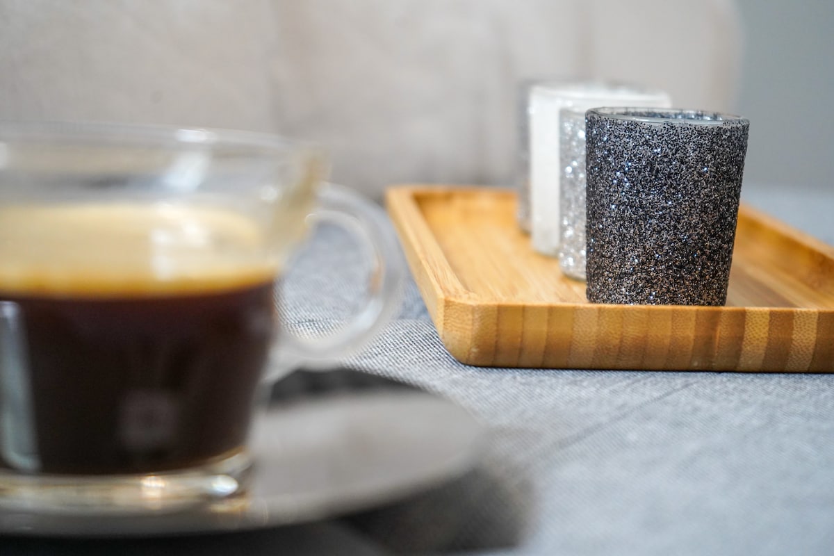 A close-up view of a glass cup filled with coffee sits in the foreground. In the background, a wooden tray holds three decorative candles with glittery silver and black finishes, complementing the neutral tones of the setting.