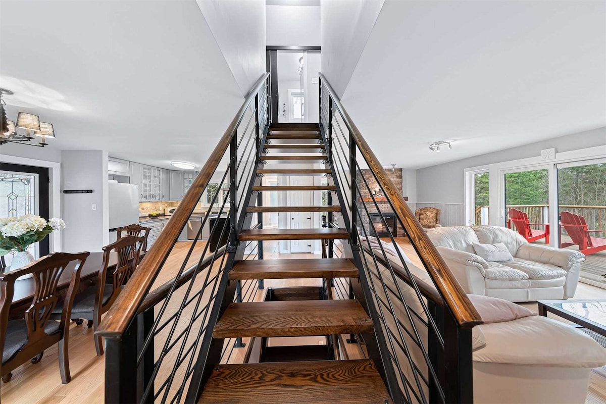 A staircase featuring wooden steps and a modern black railing leads to the upper level. Light filters down from an upper window, illuminating the adjacent open area with stylish seating and doors that open to the deck, blending indoor and outdoor living.