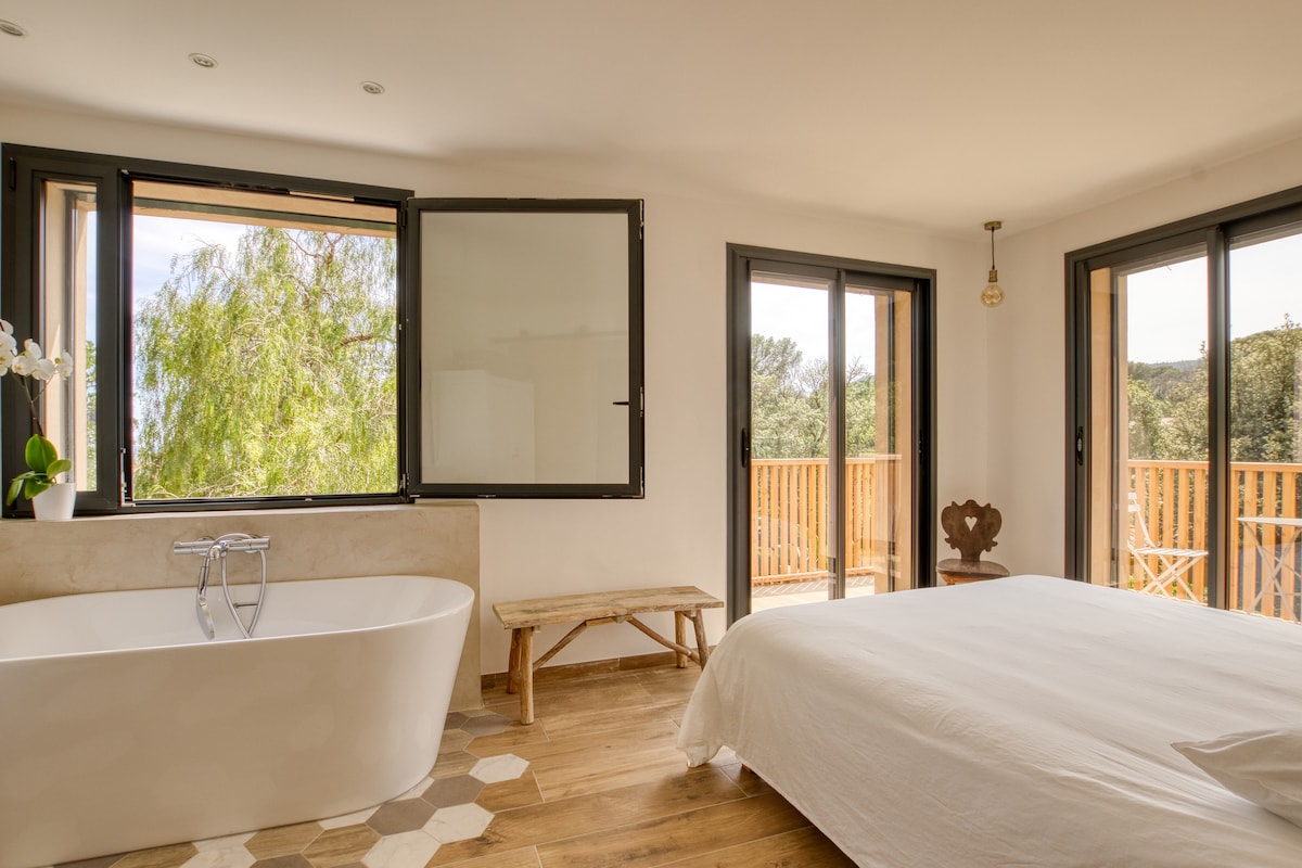 The spacious bedroom features large windows offering views of greenery and natural light. A freestanding bathtub is positioned near the windows, and a wooden bench sits beside it. A double bed is visible, with light bedding and wooden flooring enhancing the serene ambiance.