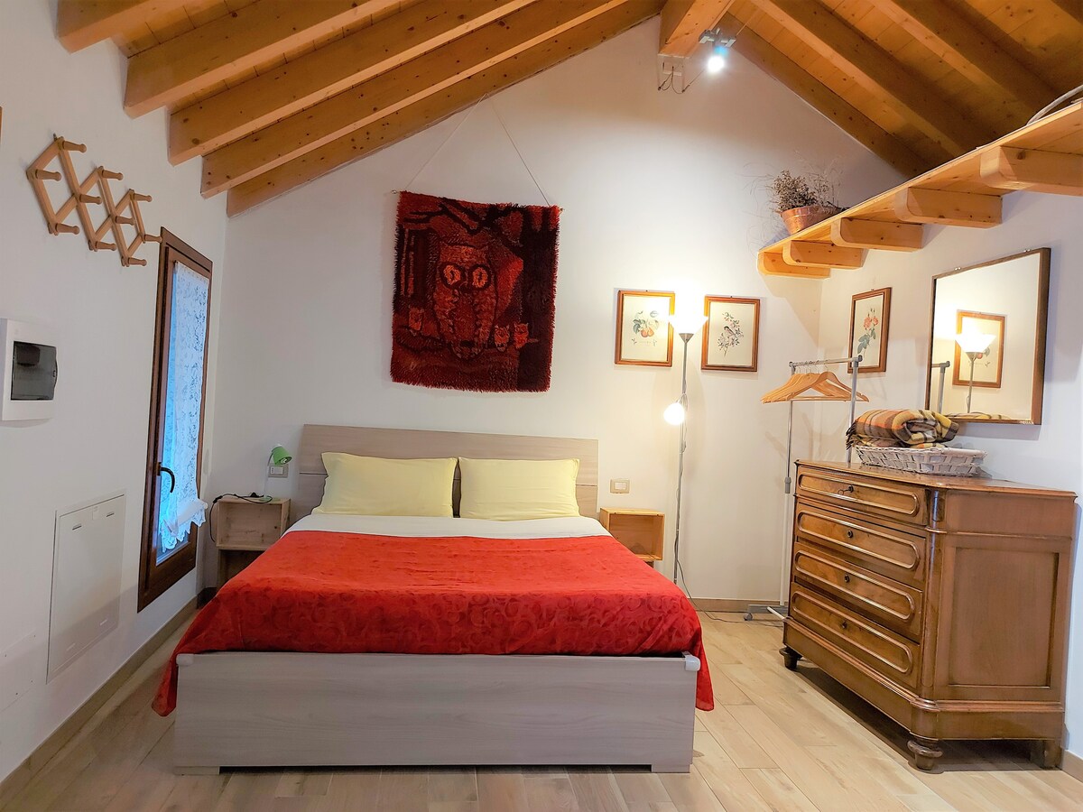 A spacious bedroom features a comfortable bed dressed in red and yellow linens beneath a wooden ceiling with exposed beams. Artwork hangs on the walls, and a wooden dresser complements a floor lamp and a clothing rack, creating a cozy ambiance.
