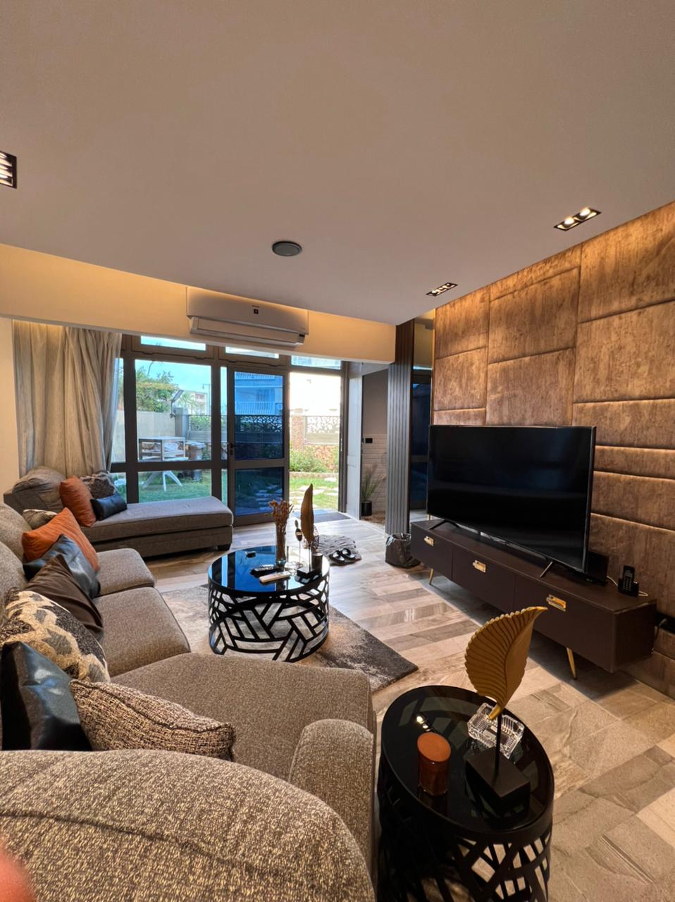 A modern living area features a comfortable gray sofa and an accent chair, complemented by decorative cushions. A sleek black TV unit displays a large screen, while a round coffee table holds stylish decor. Glass doors allow natural light and offer a view of the garden.