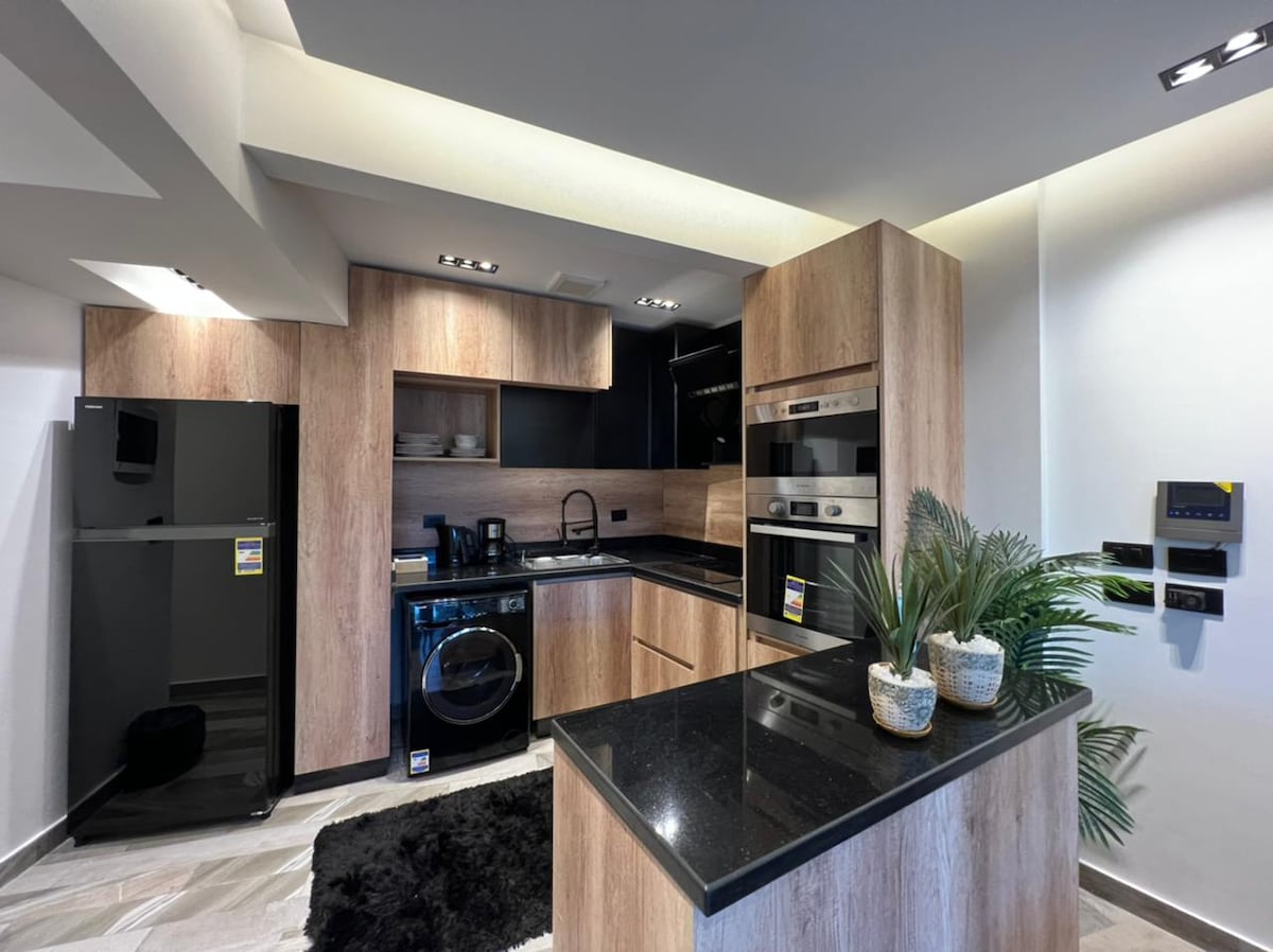 A modern kitchen features sleek wooden cabinetry and black countertops. Appliances, including a refrigerator and washer, are arranged for efficiency. Potted plants add a touch of greenery, while soft lighting highlights the clean lines and contemporary design.