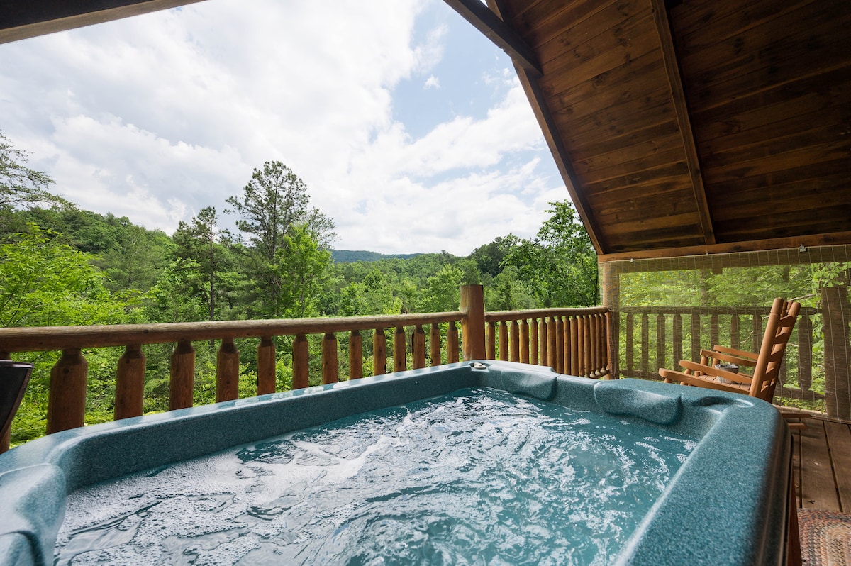 A spacious hot tub is situated on a private outdoor deck, surrounded by natural wood beams. The view beyond features lush green trees and distant hills under a partly cloudy sky, creating a serene atmosphere for relaxation.